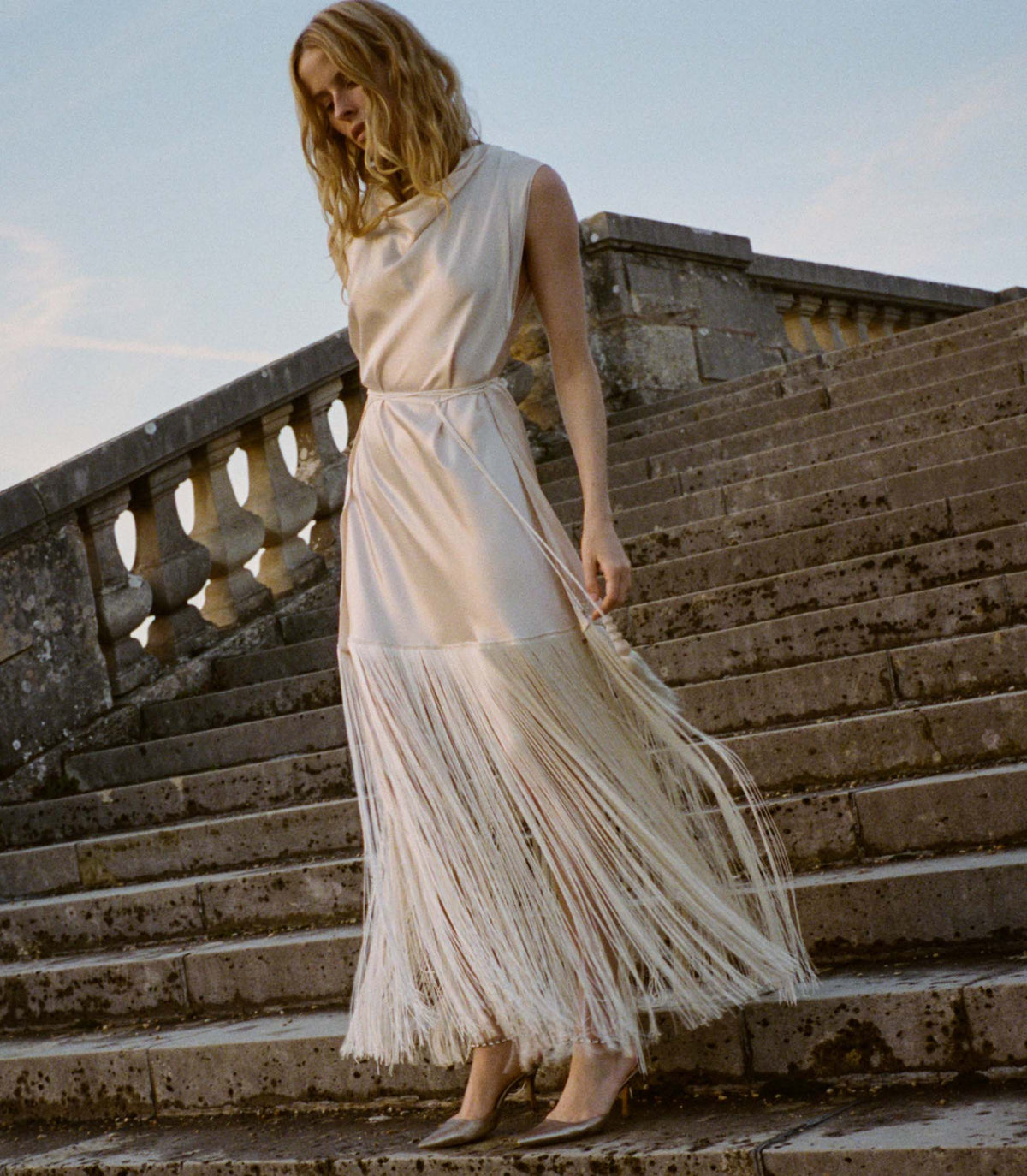 Woman in an off white flowing silk dress standing on stone steps.
