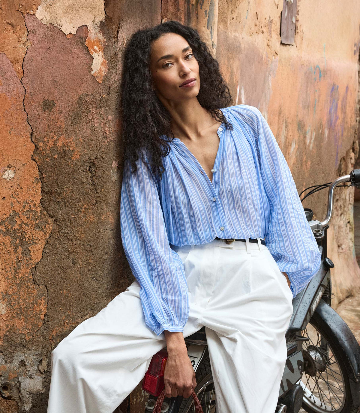 Woman in a striped blue shirt and white pants sitting on a motorcycle against a textured wall.