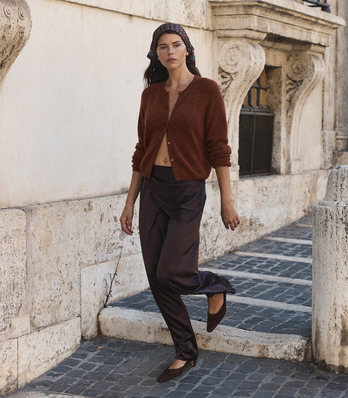 A woman wearing a rust-brown cardigan, dark brown draped trousers, and brown flats walks down a stone-paved street next to a beige stucco building.