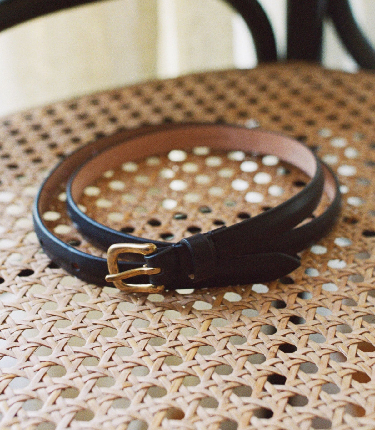 A black leather belt with a gold-tone buckle, coiled on a woven rattan chair seat.